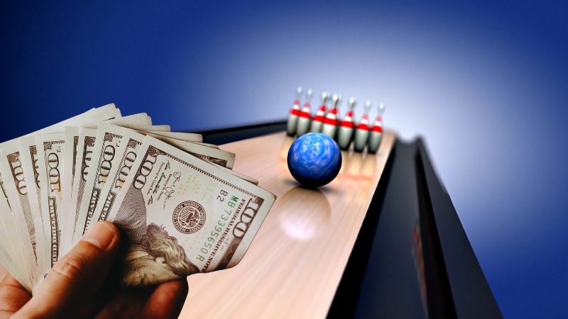 A hand holding cash with a bowling ball and pins, illustrating bowling cost